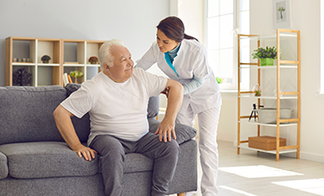Female nurse assisting happy senior patient with daily activities in nursing home. Young woman helping aged man to stand up from couch in room of modern hospital or geriatric rehabilitation center